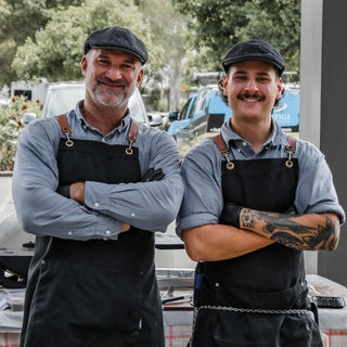Two men wearing black aprons and caps standing outdoors with trees in the background.